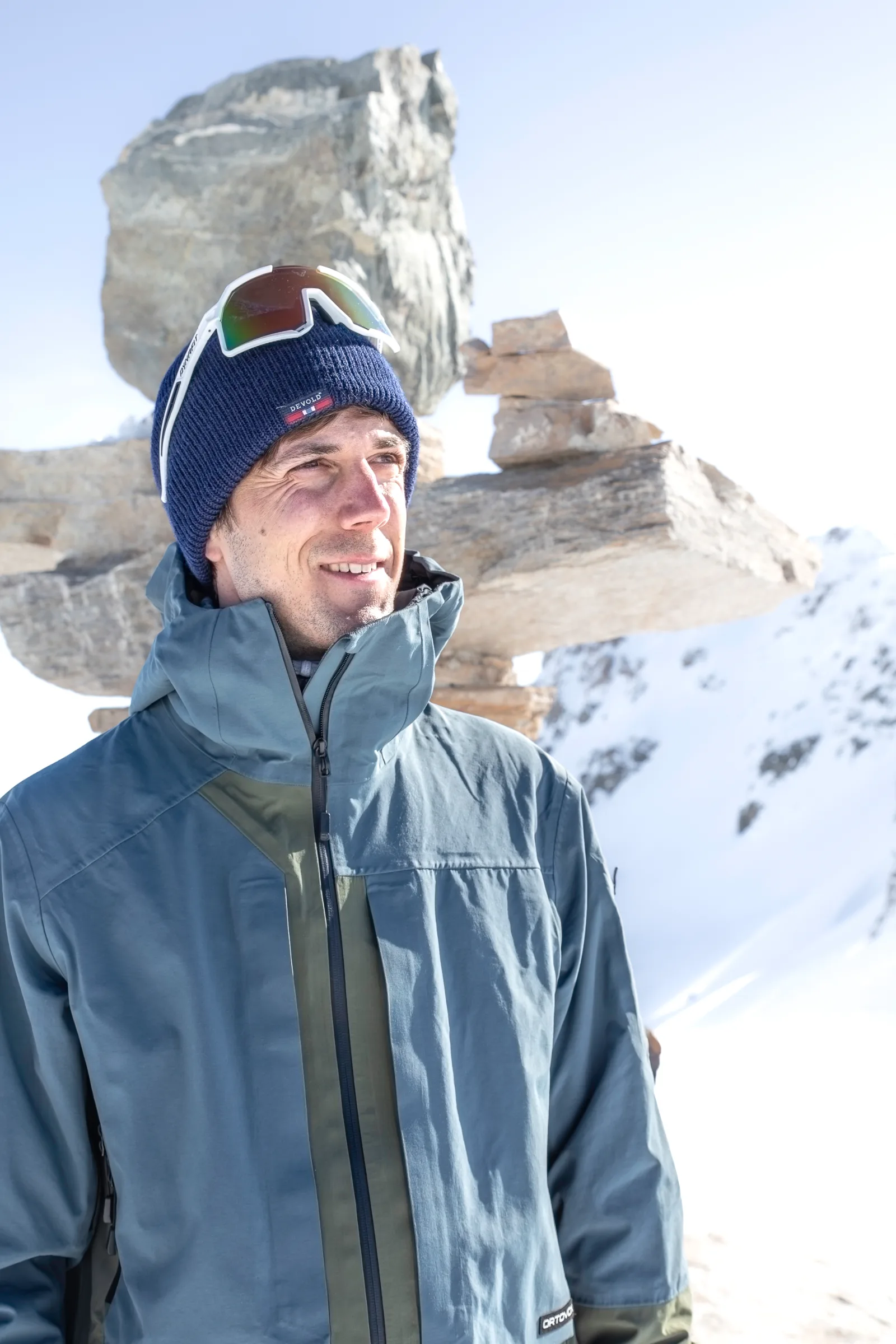 Valentin Rapp blickt mit blauer Mütze und darauf sitzender Sonnenbrille lächelnd zur Seite, vor aufgeschichteten Steinen und verschneiten Bergen.