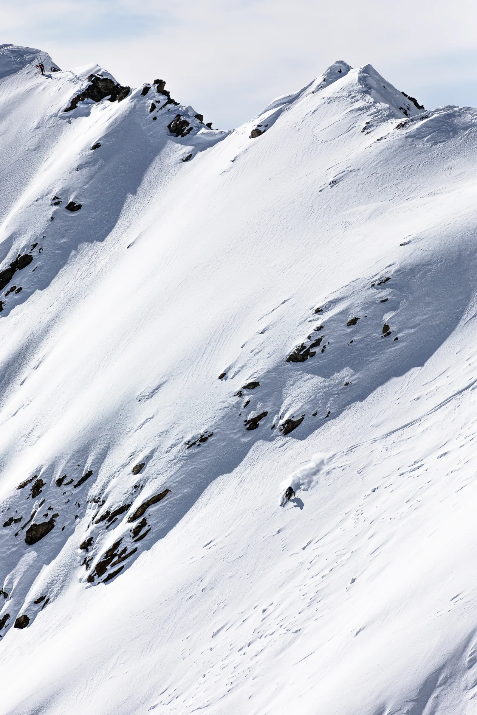 Valentin Rapp fährt einen weiten, felsdurchsetzten Steilhang bei Lenzerheide hinab und zieht eine Schneefahne hinter sich; oben am Grat steht eine weitere Person.