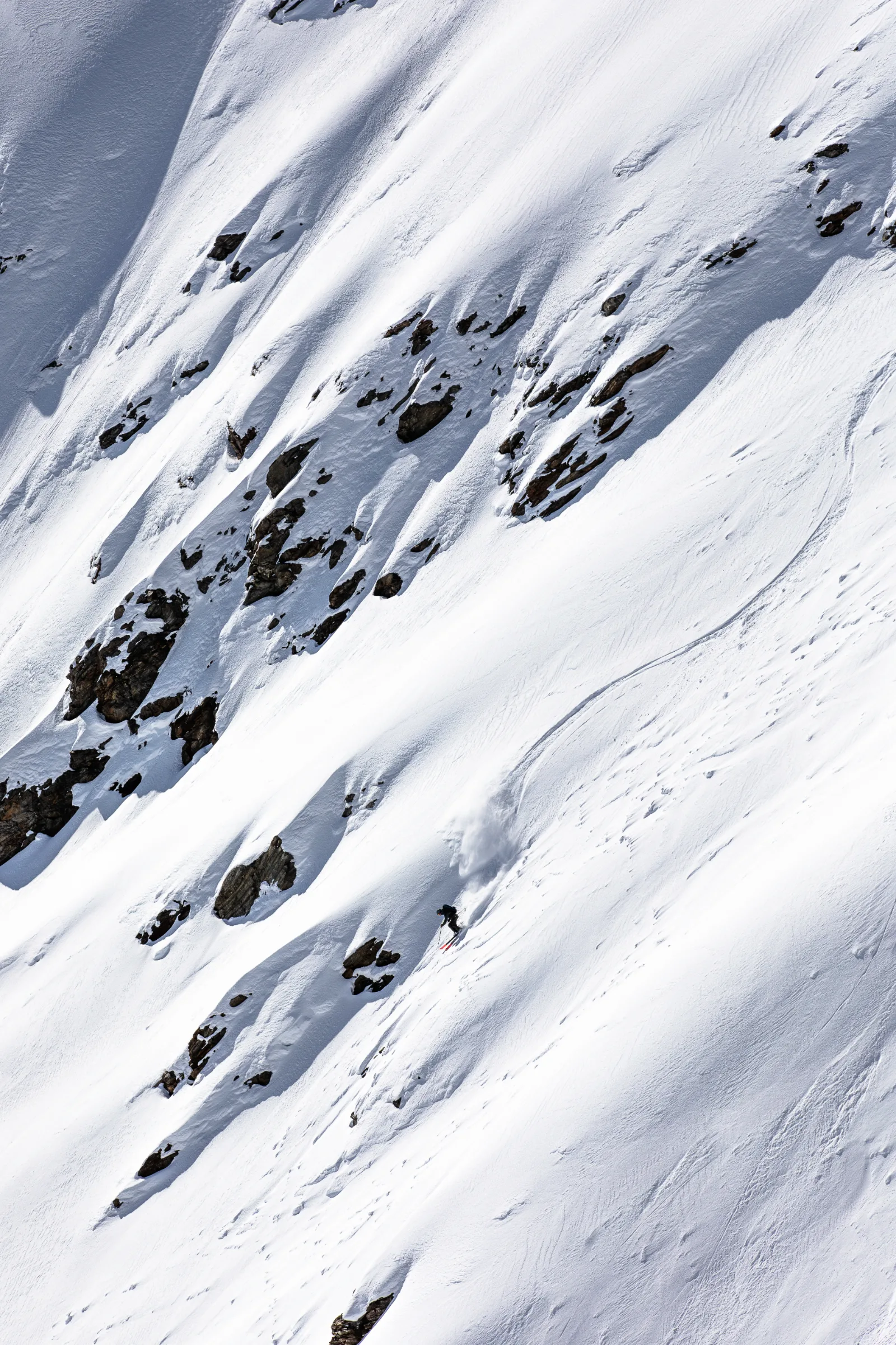 Valentin Rapp fährt neben freiliegenden Felsen einen steilen Schneehang hinab; eine geschwungene Skispur und aufgewirbelter Schnee bleiben hinter ihm zurück.
