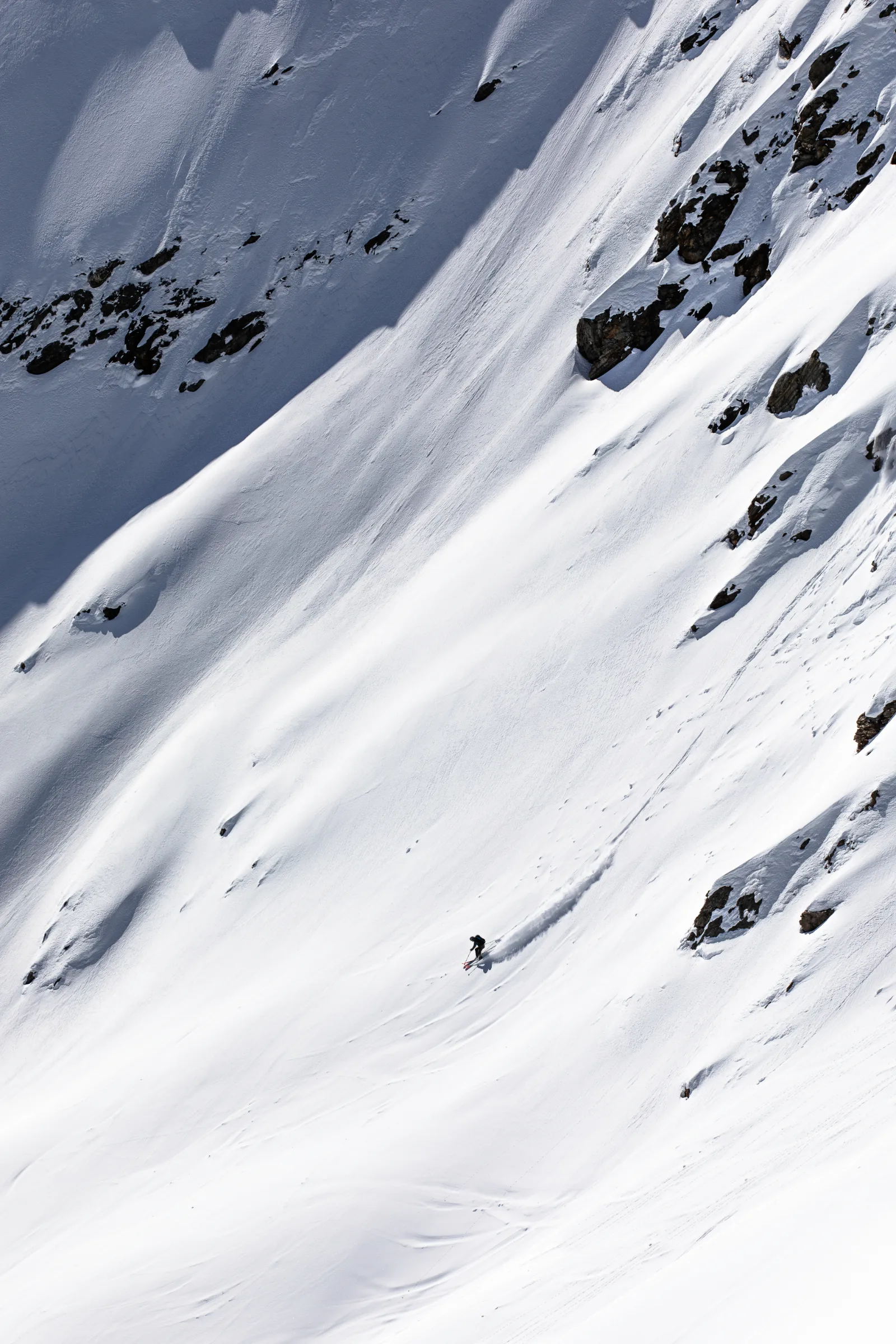 Valentin Rapp zieht einen Schwung durch einen weiten Schneehang unterhalb dunkler Felsen und wirbelt eine lange Schneefahne auf.