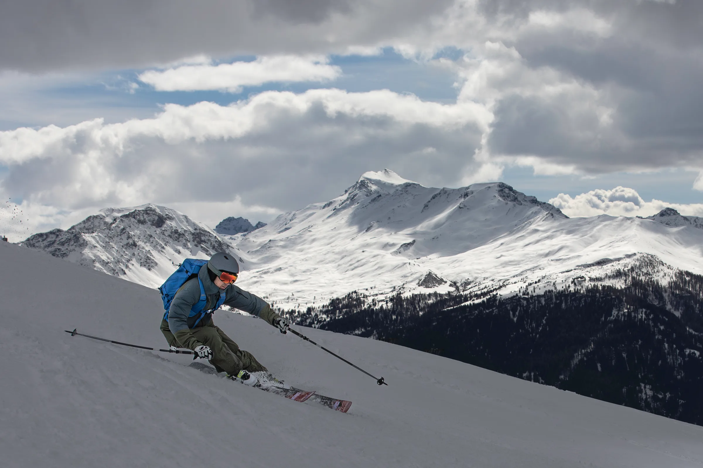 Valentin Rapp legt sich mit Helm und blauem Rucksack in einen Skischwung, vor bewaldeten Hängen und verschneiten Gipfeln bei Lenzerheide.
