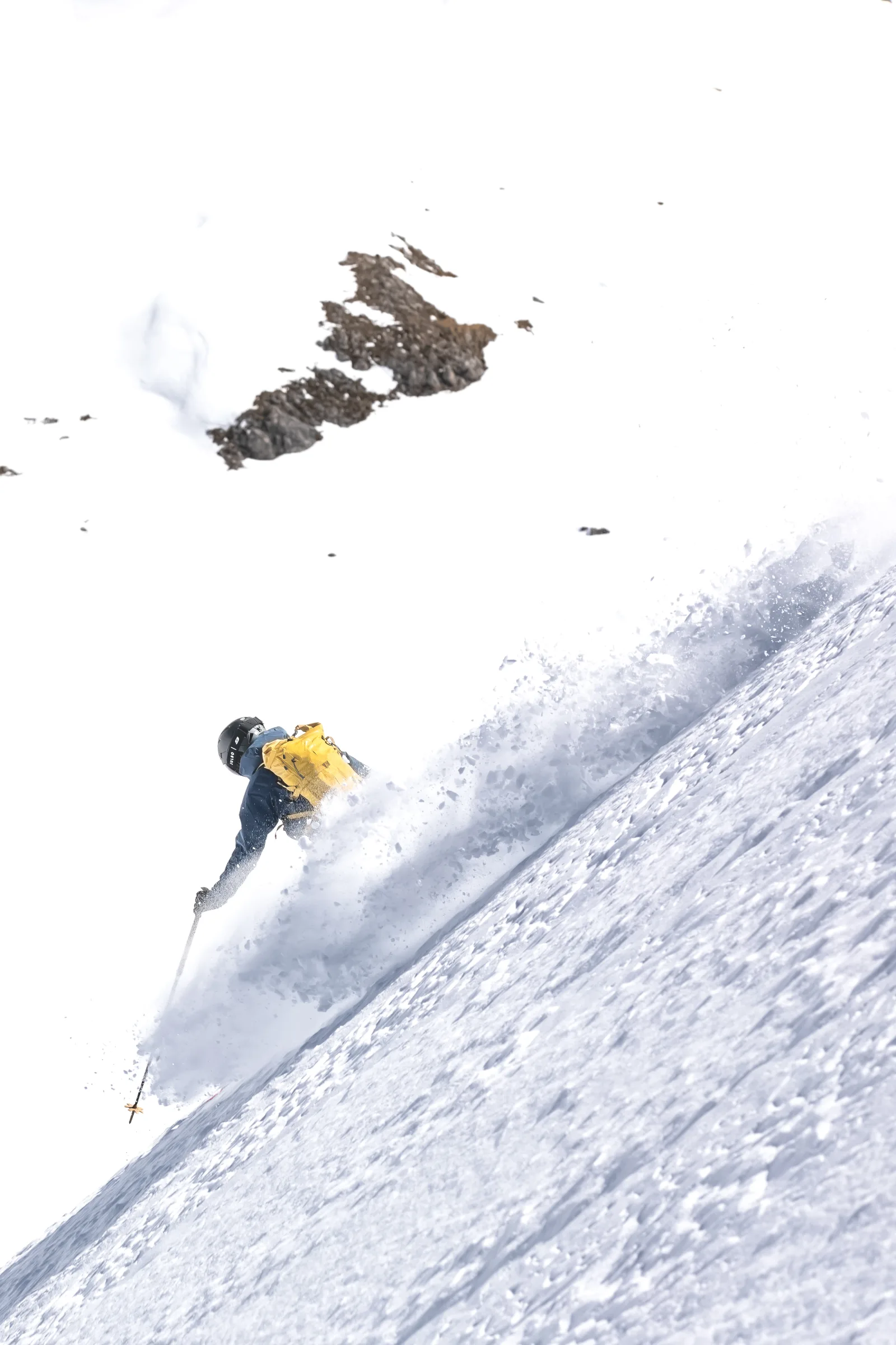 Valentin Rapp zieht mit gelbem Rucksack einen Schwung durch einen steilen Tiefschneehang unterhalb freiliegender Felsen und wirbelt eine breite Schneefahne auf.