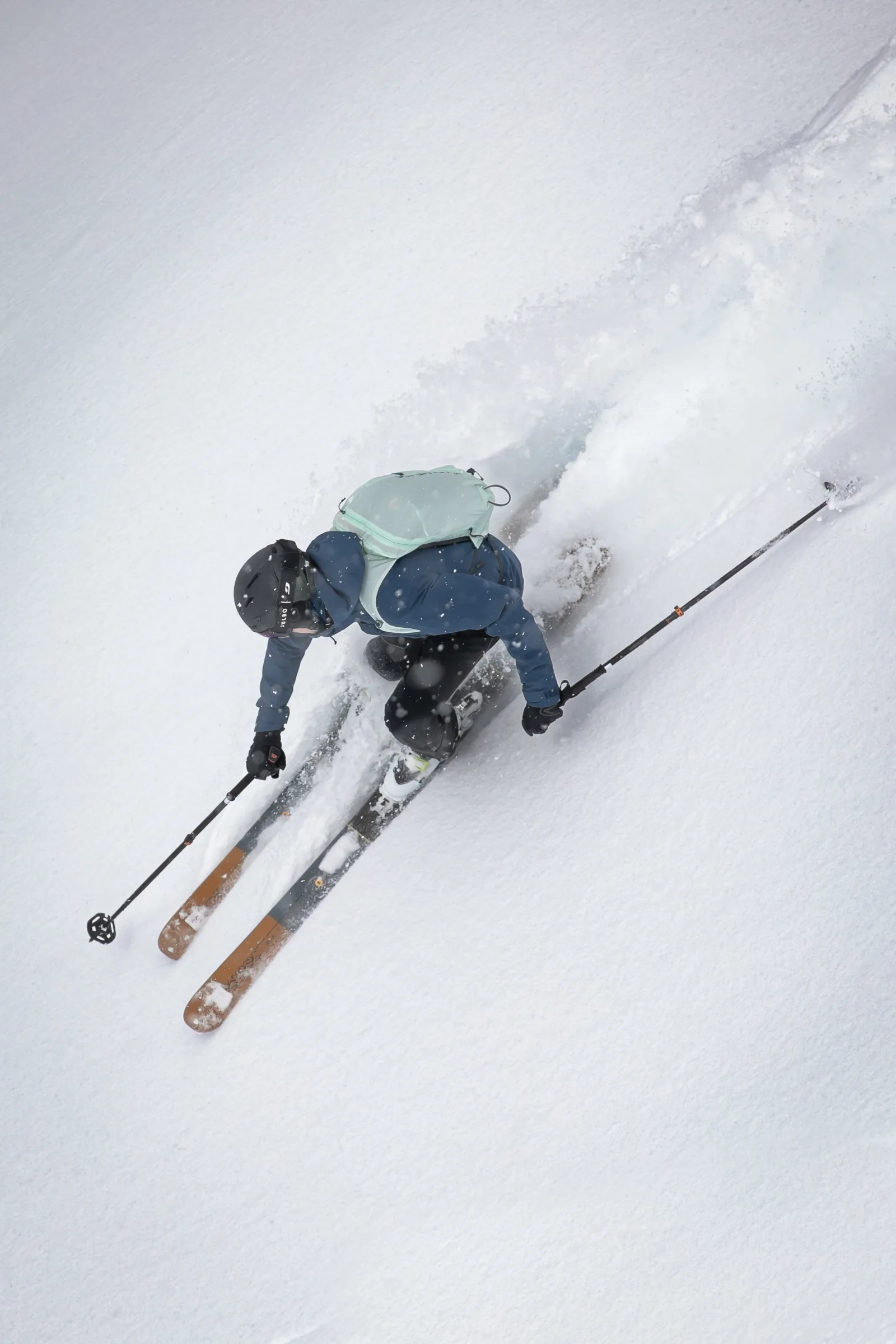 Von oben gesehen fährt ein Skifahrer mit hellgrünem Rucksack und blauer Jacke in tiefer Hocke durch Pulverschnee, die Skistöcke seitlich ausgestreckt.