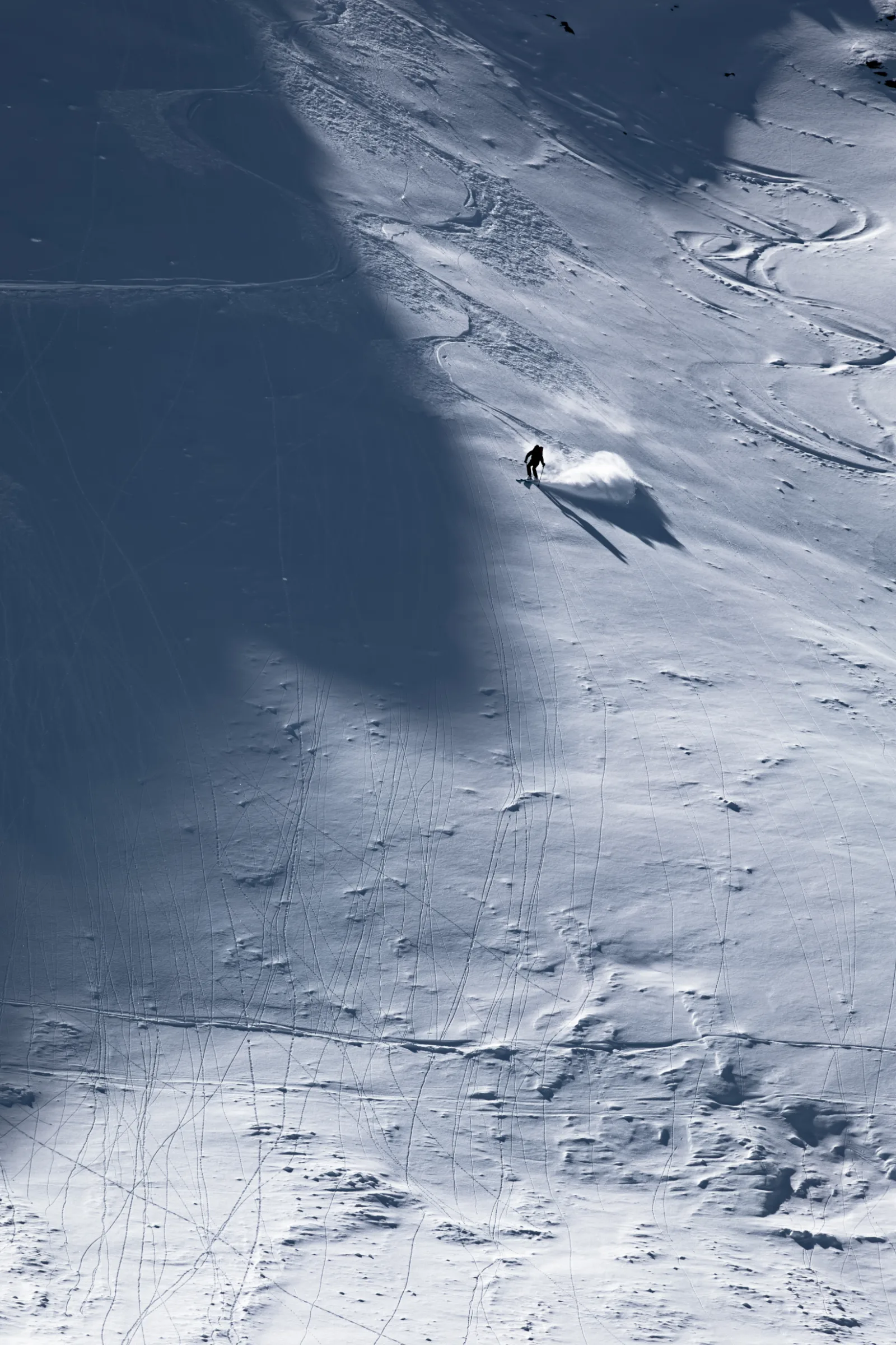 Ein einzelner Skifahrer zieht auf einem weiten, von Spuren durchzogenen Schneehang einen Schwung und wirft neben einer hellen Schneewolke einen langen Schatten.