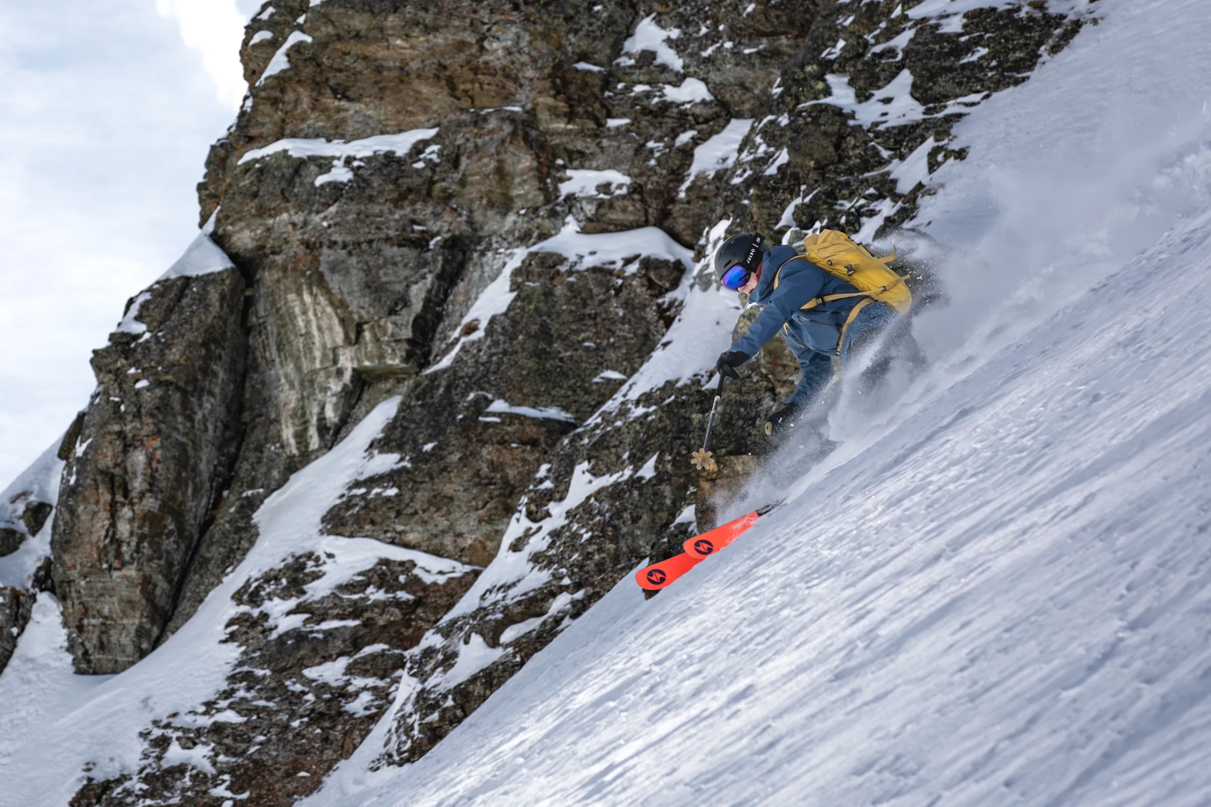 Valentin Rapp fährt mit gelbem Rucksack und leuchtend roten Skiern einen steilen Schneehang direkt neben einer zerklüfteten Felswand hinab.