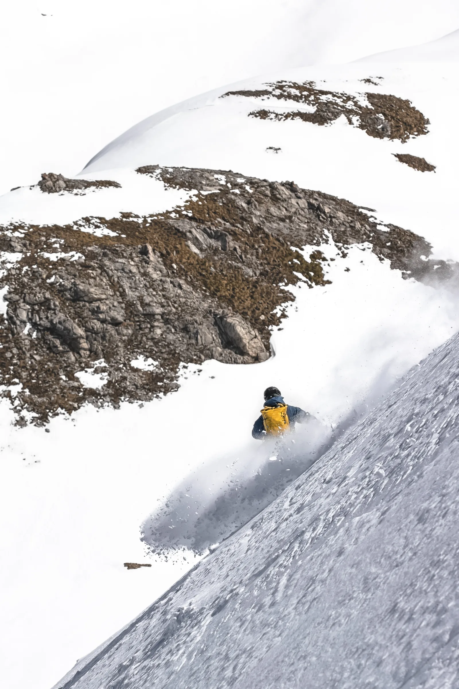 Von hinten gesehen fährt Valentin Rapp mit gelbem Rucksack durch eine große Schneewolke am steilen Hang, hinter ihm liegen schneebedeckte Felsrücken.
