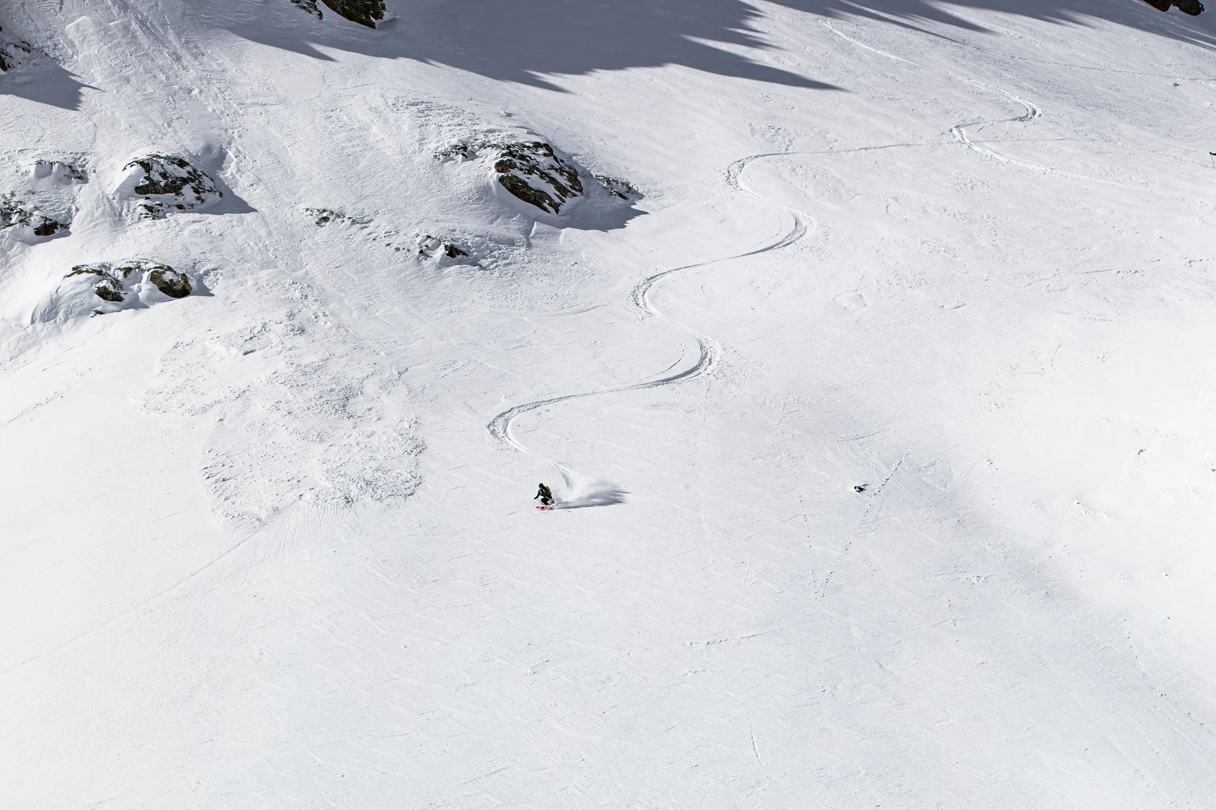 Valentin Rapp zieht auf einem weiten Schneehang in Lenzerheide einen Schwung; seine lange S-förmige Spur führt zwischen verstreuten Felsen bergauf.